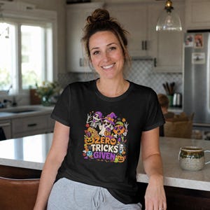 May include: A black short-sleeve t-shirt featuring a colorful graphic print of skeletons, a ghost, and pumpkins with the text "ZERO TRICKS GIVEN" in bold, stylized lettering. The design includes purple, orange, and green accents throughout.