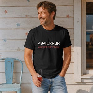 May include: A black short-sleeved t-shirt featuring the white pixelated text 404 ERROR and the red text COSTUME NOT FOUND printed on the chest. The shirt has a crew neckline and a standard fit, constructed from a solid black fabric material.