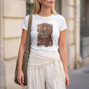 Peut inclure: Un t-shirt blanc à manches courtes avec une illustration graphique centrale représentant une personne aux longs cheveux portant un chapeau à larges bords, assise en train de lire un livre et tenant une tasse, dans un cadre ornemental sombre.