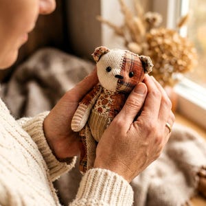 K&ouml;nnte beinhalten: Ein kleiner Stoffteddyb&auml;r aus verschiedenen Stoffflicken, darunter cremefarbener Fischgr&auml;tstoff, braunes Karomuster und ein Blumenmuster. Der B&auml;r hat schwarze Perlenaugen, eine gestickte Nase und Gliedma&szlig;en aus gestreiften und Cordstoffen.