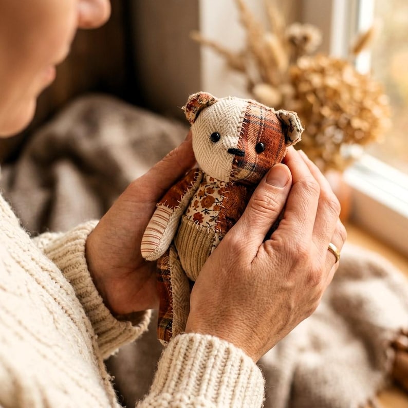 K&ouml;nnte beinhalten: Ein kleiner Stoffteddyb&auml;r aus verschiedenen Stoffflicken, darunter cremefarbener Fischgr&auml;tstoff, braunes Karomuster und ein Blumenmuster. Der B&auml;r hat schwarze Perlenaugen, eine gestickte Nase und Gliedma&szlig;en aus gestreiften und Cordstoffen.