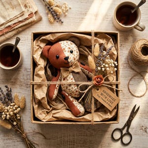 K&ouml;nnte beinhalten: Ein Stoffteddyb&auml;r mit einem Patchwork-Design aus einfarbig rostfarbenem Stoff und einem cremefarbenen Blumenmuster, der eine kleine Fliege aus Jute tr&auml;gt. Der B&auml;r liegt in einer mit braunem Papier ausgelegten Pappschachtel, die mit einer Schnur umwickelt ist.