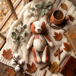 K&ouml;nnte beinhalten: Ein Patchwork-Teddyb&auml;r aus cremefarbenem und rostfarbenem Stoff mit Blumenmustern, platziert auf einer grob gestrickten Decke neben einer Keramiktasse mit Kaffee, getrocknetem Eukalyptus, Baumwollzweigen, Zimtstangen und Herbstbl&auml;ttern.