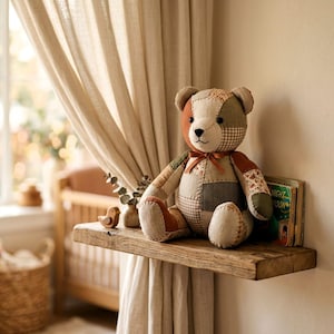 K&ouml;nnte beinhalten: Ein Stoffteddyb&auml;r aus verschiedenen Stoffflicken in Braun-, Beige- und Olivgr&uuml;nt&ouml;nen. Der B&auml;r hat ein kleines braunes Band um den Hals, dunkle Knopfaugen und eine gestickte Nase und sitzt aufrecht auf einem h&ouml;lzernen Regalbrett.