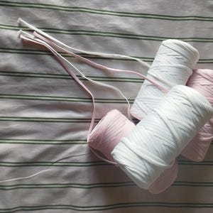 Peut inclure: Quatre bobines de cordon élastique plat, deux blanches et deux rose clair, disposées en groupe. Chaque bobine est enroulée d'une longueur continue d'élastique recouvert de tissu, avec des extrémités libres s'étendant depuis le centre.