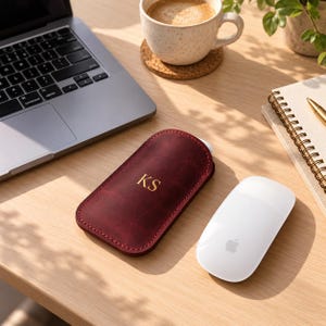 May include: A deep burgundy leather sleeve for a computer mouse, featuring the gold embossed initials KS on the front. The sleeve has visible stitching along the edges and is positioned on a wooden desk next to a laptop, a white mouse, and a notebook.