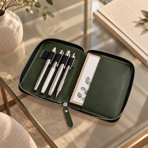 Puede incluir: Un estuche organizador con cremallera en color verde oscuro que cuenta con cuatro bucles elásticos para bolígrafos plateados y blancos, y un bolsillo lateral con un bloc de notas con una ilustración de hojas verdes. Incluye tirador a juego.
