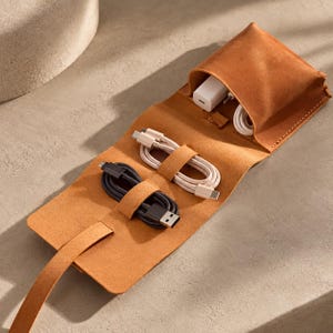 Könnte beinhalten: Ein beigefarbener Leder-Organizer zum Aufrollen mit einer oberen Tasche, die ein weißes Netzteil und ein weißes Kabel enthält, sowie zwei zusätzlichen elastischen Schlaufen für ein weißes USB-C-Kabel und ein schwarzes USB-A-auf-USB-C-Kabel.