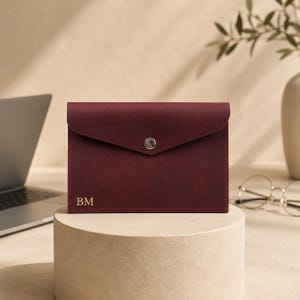 May include: A rectangular burgundy leather envelope pouch featuring a pointed flap with a single silver-toned metal snap button closure. The bottom left corner of the pouch is embossed with the gold-coloured capital letters BM.