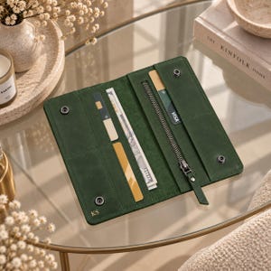 Può includere: Un portafoglio lungo in pelle verde scuro con chiusura a bottone automatico, scomparti per carte, una tasca con cerniera e una banconota visibile. Il portafoglio presenta le iniziali KS impresse in oro nell'angolo in basso a sinistra.