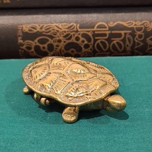 Peut inclure: Une petite figurine de tortue en métal doré avec un motif de carapace texturé et détaillé. La tortue est représentée dans une position basse, en train de ramper, avec la tête étendue vers l'avant et quatre pattes visibles, offrant une surface polie.