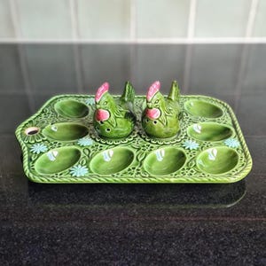 May include: A green ceramic egg tray featuring ten oval indentations and a decorative floral pattern. Two matching green chicken-shaped salt and pepper shakers with pink combs and wattles are positioned in the center of the rectangular tray.