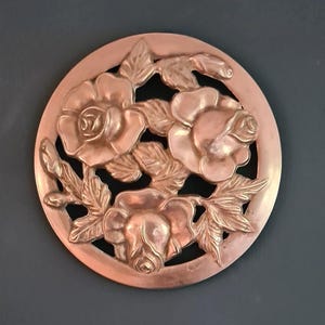 May include: A circular copper-toned metal wall plaque featuring a relief design of three blooming roses surrounded by detailed leaves. The floral arrangement is set within a solid circular frame, creating an openwork effect with dark negative space.