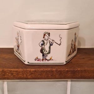 Może przedstawiać: Białe, sześciokątne pudełko ceramiczne z pokrywką, ozdobione ręcznie malowanymi ilustracjami postaci w historycznych strojach. Środkowy panel przedstawia osobę w tunice palącą długą fajkę, a panele boczne ukazują inne postacie w plenerowych scenach.