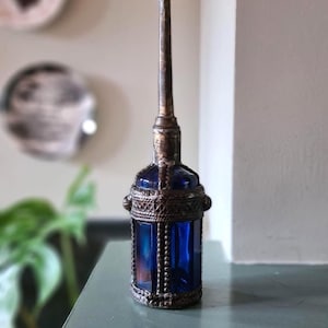 Op de afbeelding: Een vintage oliefles van kobaltblauw glas met een hoge, slanke metalen tuit en een decoratieve, getextureerde metalen kooi. Het cilindrische glazen lichaam is gevat in een donker, verweerd metalen frame met gedetailleerde versieringen.