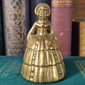 Könnte beinhalten: Eine Messingglocke in Form einer Figur mit Haube und einem gestuften, bodenlangen Kleid. Die Figur hat die Hände in die Hüften gestützt und der Rock weist vertikale Reihen erhabener kreisförmiger Details auf. Das Metall hat ein goldenes Finish.