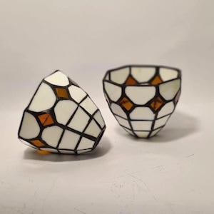Pode incluir: Dois suportes para velas em vitral com um padrão geométrico de painéis de vidro branco opaco intercalados com pequenos detalhes de vidro cor de âmbar em forma de diamante, unidos por linhas de solda de metal escuro em formato de taça.
