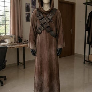 以下が含まれることがあります： 質感のある生地で作られたフルレングスの茶色のフード付きローブ。複数の小さなポーチと金属製のリングが付いたダークブラウンのレザー製チェストハーネスを着用しています。マネキンには黒い手袋と黒いブーツが組み合わされています。