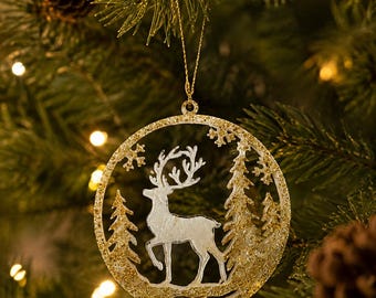 May include: A circular Christmas tree ornament featuring a white silhouette of a standing deer surrounded by pine trees. The frame is covered in gold glitter, and the ornament hangs from a thin gold string, creating a festive holiday decoration.