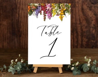 May include: A white table number card with the word "Table" and the number "1" in black script. The top of the card is decorated with a colourful floral arrangement. The card is displayed on a small wooden easel.