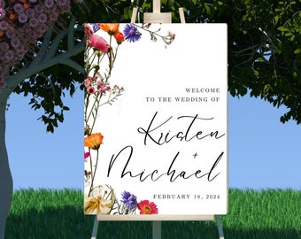 Op de afbeelding: Een wit huwelijksbord met een bloemige rand en de tekst "Welkom op de bruiloft van Kirsten + Michael 18 februari 2024" op een houten ezel in een grasveld.