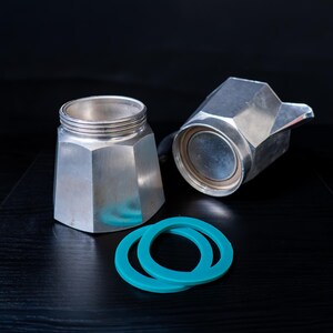 May include: A disassembled silver-toned metal moka pot featuring an octagonal base with threaded top, a separate upper chamber with a spout, and two circular teal-colored silicone gaskets positioned in front of the coffee maker components.