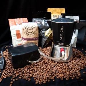 May include: A black Grosche Milano Induction moka pot with a silver base, connected by a clear tube to a black rectangular electric pump with a power cord and switch, all resting on a bed of roasted coffee beans with coffee bags in the background.