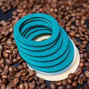 May include: A stack of circular, teal-colored silicone gaskets with a smooth, flat surface. The stack includes several identical teal rings resting on top of a single, larger, solid off-white circular disc base, creating a tiered, cylindrical shape.