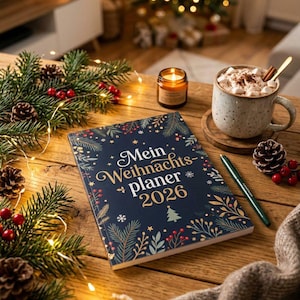 Könnte beinhalten: Ein dunkelblauer Planer mit dem Text Mein Weihnachts-planer 2026, verziert mit goldenen und grünen botanischen Illustrationen von Tannenzweigen und Beeren. Ein grüner Stift, eine Tasse heiße Schokolade mit Marshmallows und eine Kerze auf einem Holztisch.