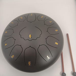 May include: A dark gray steel tongue drum with a circular shape and multiple petal-shaped cutouts. Each cutout is marked with a gold-colored number sticker. Two wooden mallets with black rubber tips are positioned to the right of the drum.