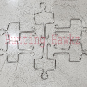 Puede incluir: Seis ganchos de alambre metálico de color plateado dispuestos en un patrón simétrico. Cada gancho presenta un marco rectangular, un bucle circular en un extremo y dos puntas curvas en el otro. El texto Hunting Hawkz aparece en color rojo.