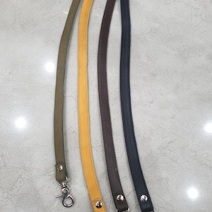 May include: Four long, narrow bag straps in olive green, mustard yellow, dark brown, and black. Each strap features a textured surface, silver-toned metal rivets at the ends, and a silver-toned swivel lobster clasp attached to each end for secure fastening.