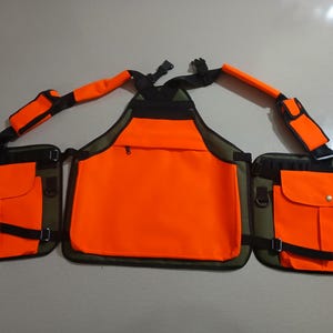 Może przedstawiać: Kamizelka typu chest rig w kolorze pomarańczowym i oliwkowym, wyposażona w dużą kieszeń środkową, dwie kieszenie boczne z klapkami oraz regulowane czarne szelki z wyściełanymi sekcjami. Konstrukcja zawiera liczne klamry i paski.