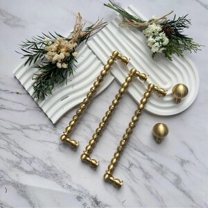 May include: Three gold-toned metal cabinet pulls with a beaded texture and two matching round knobs. The hardware is arranged alongside two white, arched, ribbed decorative trays, each adorned with a small bouquet of dried white flowers and greenery.
