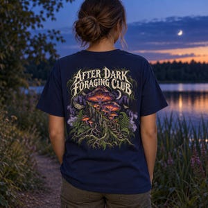 May include: A navy blue t-shirt featuring a graphic on the back with the text "AFTER DARK FORAGING CLUB" in a stylized font. The illustration depicts glowing purple and orange mushrooms growing on a tree stump, surrounded by ferns and moths.
