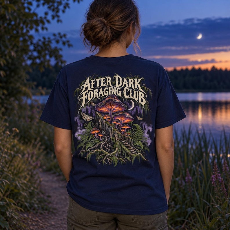 May include: A navy blue t-shirt featuring a graphic on the back with the text "AFTER DARK FORAGING CLUB" in a stylized font. The illustration depicts glowing purple and orange mushrooms growing on a tree stump, surrounded by ferns and moths.