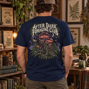 May include: A navy blue t-shirt featuring a graphic on the back with the text "AFTER DARK FORAGING CLUB" above an illustration of glowing mushrooms, ferns, tree roots, and moths in shades of purple, orange, and green against a dark background.