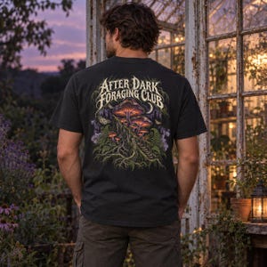 May include: A black short-sleeve t-shirt featuring a graphic on the back with the text "AFTER DARK FORAGING CLUB" in a stylized font. The illustration depicts glowing purple and orange mushrooms, green ferns, tree roots, and moths.