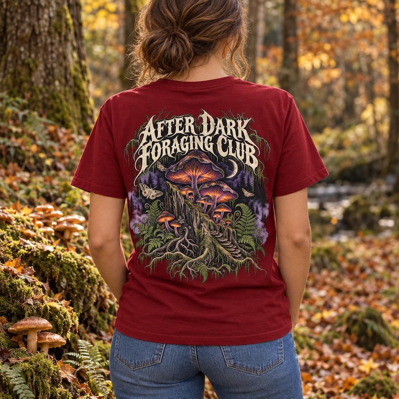 May include: A maroon short-sleeve t-shirt featuring a graphic on the back with the text "AFTER DARK FORAGING CLUB" above an illustration of glowing mushrooms on a tree stump, surrounded by ferns, moths, and a crescent moon in a forest setting.