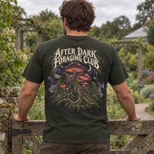 May include: A dark olive green t-shirt featuring a back graphic of glowing purple and orange mushrooms growing on a tree stump, surrounded by ferns and moths. The text "AFTER DARK FORAGING CLUB" is printed in a stylized white font above the art.
