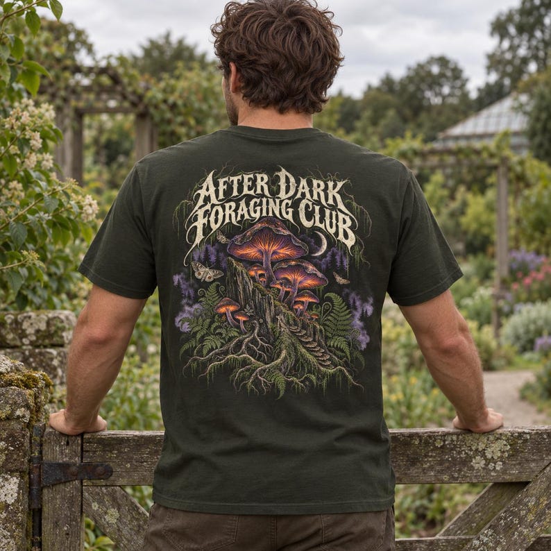 May include: A dark olive green t-shirt featuring a back graphic of glowing purple and orange mushrooms growing on a tree stump, surrounded by ferns and moths. The text "AFTER DARK FORAGING CLUB" is printed in a stylized white font above the art.