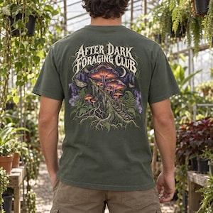 May include: An olive green t-shirt featuring a back graphic of glowing purple and orange mushrooms on a mossy log, surrounded by ferns and moths. The text "AFTER DARK FORAGING CLUB" is printed in a stylized white font above the illustration.