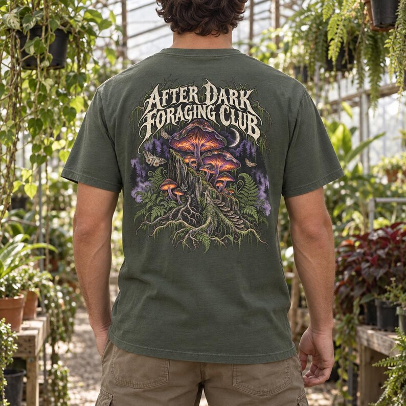 May include: An olive green t-shirt featuring a back graphic of glowing purple and orange mushrooms on a mossy log, surrounded by ferns and moths. The text "AFTER DARK FORAGING CLUB" is printed in a stylized white font above the illustration.