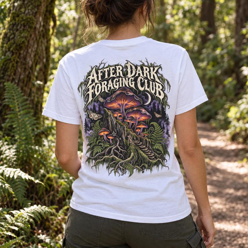 May include: A white short-sleeve t-shirt featuring a detailed graphic on the back. The design displays glowing purple and orange mushrooms, ferns, and moths with the text "AFTER DARK FORAGING CLUB" in a stylized font above the forest scene.