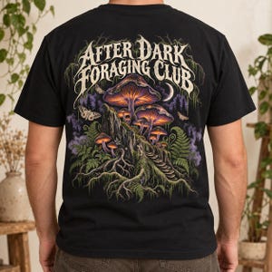 May include: A black short-sleeve t-shirt featuring a graphic on the back with the text "AFTER DARK FORAGING CLUB" in a stylized font. The design depicts glowing purple and orange mushrooms on a rocky outcrop, ferns, moths, and a crescent moon.