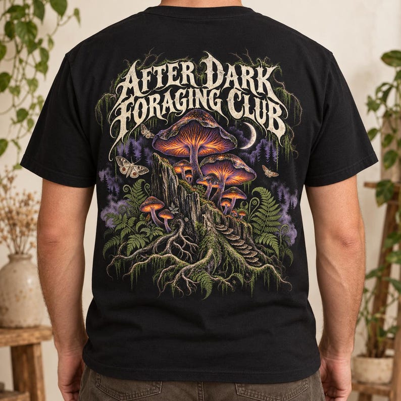 May include: A black short-sleeve t-shirt featuring a graphic on the back with the text "AFTER DARK FORAGING CLUB" in a stylized font. The design depicts glowing purple and orange mushrooms on a rocky outcrop, ferns, moths, and a crescent moon.