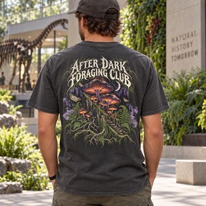 May include: A charcoal gray short-sleeve t-shirt featuring a back graphic of glowing mushrooms, ferns, and moths. The text "AFTER DARK FORAGING CLUB" is printed in a stylized white font above the illustration, which includes purple smoke accents.
