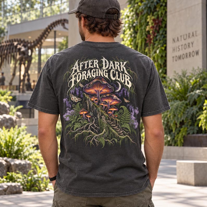 May include: A charcoal gray short-sleeve t-shirt featuring a back graphic of glowing mushrooms, ferns, and moths. The text "AFTER DARK FORAGING CLUB" is printed in a stylized white font above the illustration, which includes purple smoke accents.