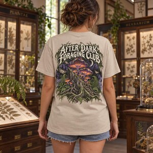 May include: A beige short-sleeve t-shirt featuring a graphic on the back with the text After Dark Foraging Club. The illustration depicts glowing purple and orange mushrooms growing on a tree stump, surrounded by green ferns and moths.