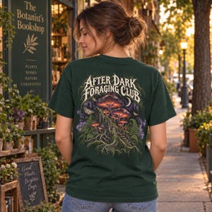 May include: A dark green t-shirt featuring a graphic on the back with the text After Dark Foraging Club. The illustration depicts glowing purple and orange mushrooms growing on a mossy log, surrounded by ferns, moths, and a crescent moon.